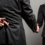 Business partners shaking hands with one of them holding fingers crossed behind back.