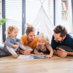 Young family with two small children indoors in bedroom reading a book.