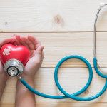 Young child kid holding red love heart hand exercise ball with bandage, MD medical doctor physician's stethoscope on light white wood background: Nursing children day care healthcare support concept