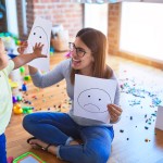 Young beautiful psicologist and toddler doing therapy using emoji emotions at kindergarten