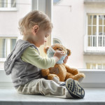 Child in home quarantine playing at the window with his sick teddy bear wearing a medical mask against viruses during coronavirus and flu outbreak. Children and illness COVID-2019 disease concept