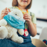 Soft toy bear with protective medical mask in child hands. Child playing doctor with soft toy.